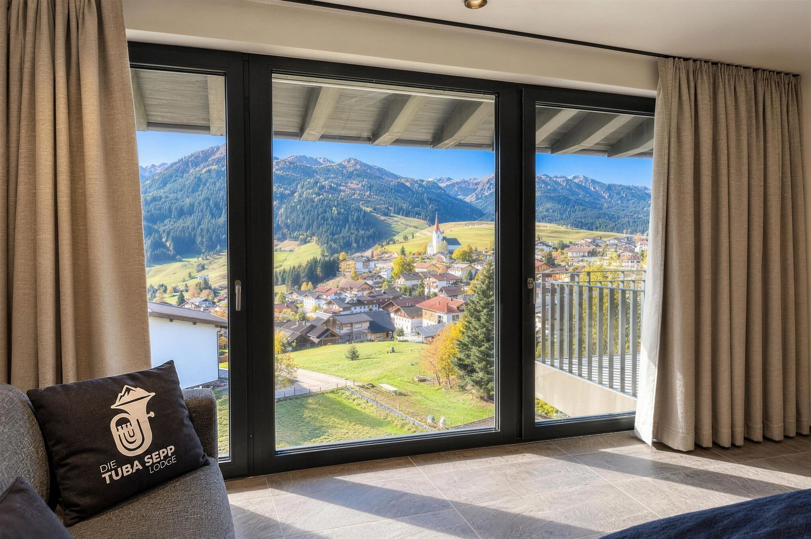 Tuba-Sepp-Kissen auf dem Sofa vor der Fensterfront mit Blick auf das Dorf, die Kirche und die Berge